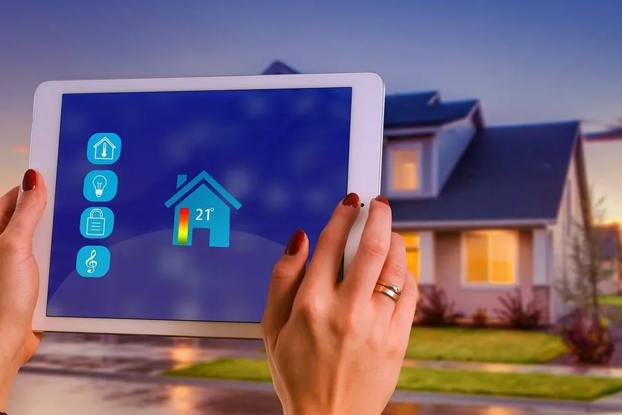Mãos segurando um tablet com aplicativo de casa inteligente, com uma casa iluminada ao fundo